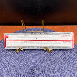 May include: A vintage Gisholt Time Computer slide rule, Model M, with red and white markings. The slide rule is displayed on a blue woven mat and held by a gold-colored metal stand. The price is $3.00.