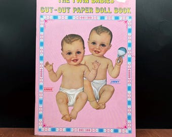 May include: A vintage paper doll book titled "The Twin Babies Cut-Out Paper Doll Book." The cover is pink with a border and features illustrations of two babies, one holding a rattle. The babies are wearing diapers, and the names "Annie" and "Jimmy" are printed below them.