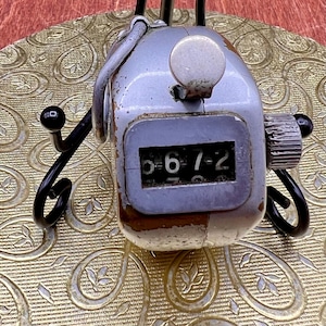 May include: A silver-toned handheld mechanical tally counter featuring a prominent push-button actuator on top, a side-mounted reset knob, and a digital display window showing the numerical sequence 6-7-2 against a black background.