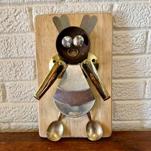 May include: A wall-mounted art piece featuring a figure constructed from repurposed metal kitchen utensils attached to a rectangular wooden board. The figure includes a dark bowl head, silver spoon ears, brass handles for arms, and two small brass spoons as feet.