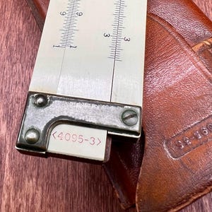 May include: Close-up of a vintage slide rule with a cream-colored scale and metal frame. The scale has black markings and numbers. The slide rule is next to a brown leather case with stamped text.