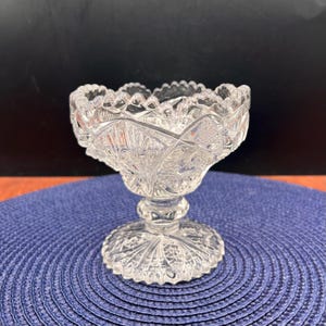 May include: A clear, cut-glass pedestal bowl with a scalloped rim. The bowl features intricate geometric and floral patterns, and sits on a round base. The bowl is likely used for serving desserts or displaying small items.