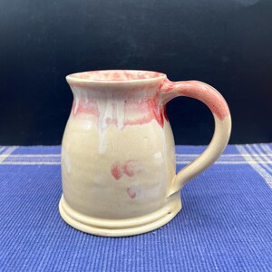 May include: A handmade ceramic mug with a cream-colored base and a red glaze dripping from the rim and handle. The mug has a rounded body and a sturdy handle, perfect for enjoying your favorite beverage.
