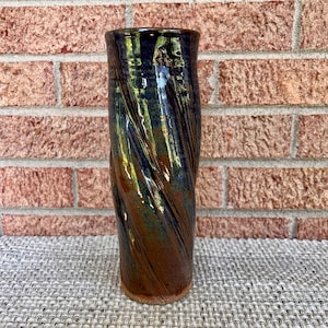 May include: A tall, cylindrical ceramic vase featuring a glossy, multi-toned glaze. The surface displays a swirling, textured pattern with deep shades of navy blue, metallic gold, and earthy brown, creating a dynamic, reflective appearance.