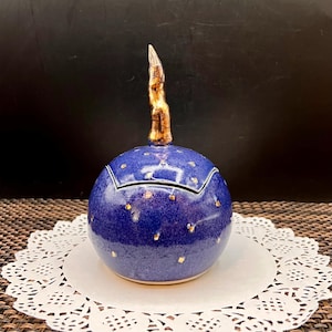 May include: A spherical ceramic jar with a deep blue glaze and scattered gold-colored flecks. The lid features a prominent, textured, metallic gold-toned finial that tapers to a point, contrasting with the smooth, rounded body of the vessel.