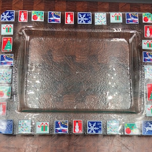 May include: A rectangular clear glass tray featuring a decorative border of colorful vintage-style Christmas postage stamps. The stamps display various holiday imagery including snowmen, snowflakes, Christmas trees, gifts, and ornaments.