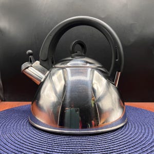 May include: A stainless steel stovetop kettle with a black handle and spout. The kettle has a shiny, reflective surface and a small black knob on the lid. It sits on a blue woven mat.