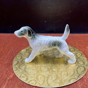 May include: A ceramic figurine of a dog, predominantly white with gray markings on its ears, back, and tail. The dog is posed in a walking stance, set on a gold-colored, patterned base. The figurine is likely a decorative item.