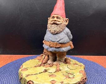 Könnte beinhalten: Eine Zwergenfigur mit einer hohen roten Zipfelmütze, einem strukturierten braunen Bart und einer blauen Tunika mit braunem Gürtel. Die Figur steht auf einem runden Sockel, der mit geformten grünen Blättern und kleinen braunen Eicheln verziert ist.