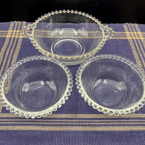 May include: A set of three clear glass serving dishes. The set includes two small round bowls and one larger oval bowl with handles. Each piece has a beaded edge.