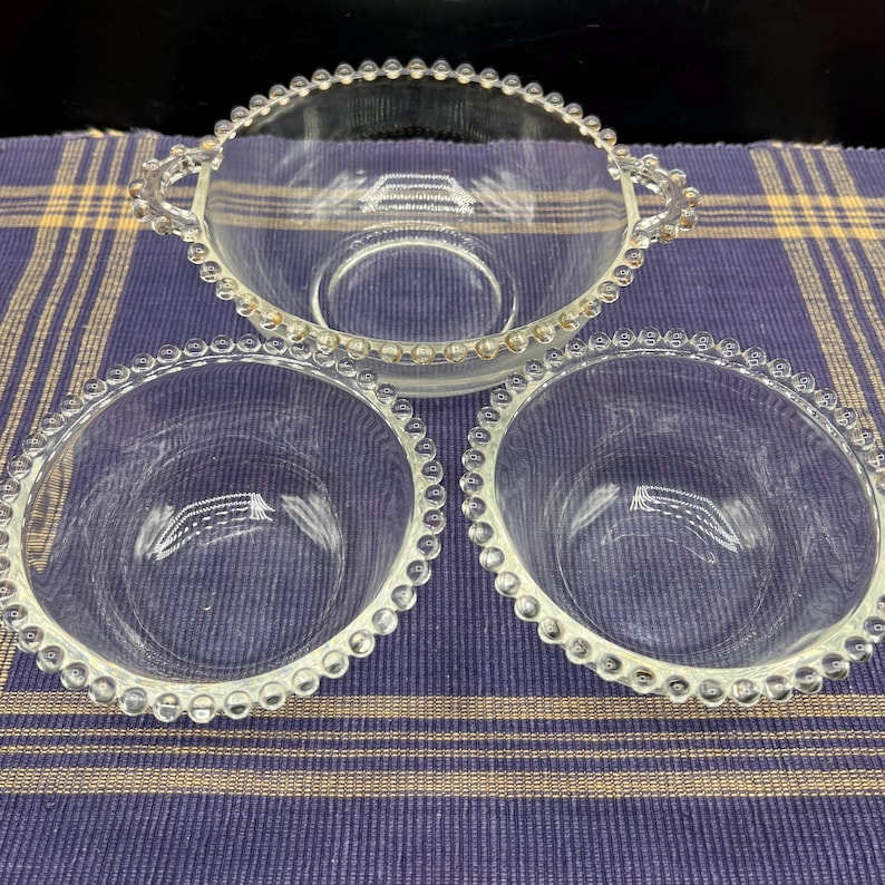 May include: A set of three clear glass serving dishes. The set includes two small round bowls and one larger oval bowl with handles. Each piece has a beaded edge.