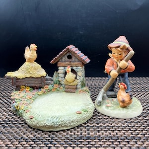 May include: Two ceramic figurines depicting a farm scene. One features a boy with a rake and a chicken, the other has a chicken coop and a chicken on a hay bale. The figurines are painted with detailed colors.