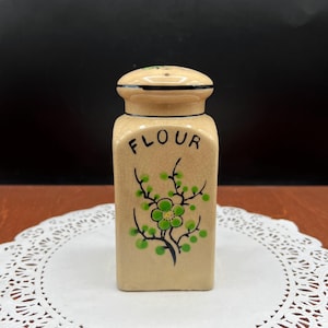 May include: A beige ceramic flour container with a lid. The word "FLOUR" is written in black on the front. A green floral design with black stems decorates the front. The container is on a white doily.