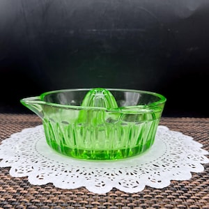 May include: A vintage, light green glass citrus juicer. The juicer has a ribbed bowl, a reamer, and a spout for pouring. It sits on a white, lacy paper doily. The background is dark.