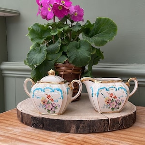 May include: A white sugar bowl and creamer set with a floral design and blue trim. The set is on a wooden slice and a potted plant with pink flowers is in the background.