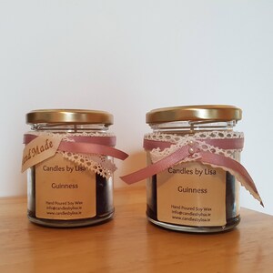 Op de afbeelding: Twee glazen potten met gouden deksels en roze lintaccenten. Elke pot bevat een donkerbruine kaars en een label met de tekst "Candles by Lisa" en "Guinness".