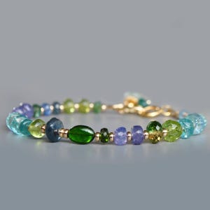 Peut inclure: Un bracelet de perles avec des pierres précieuses colorées. Le bracelet comprend des pierres bleues, vertes et violettes, avec des accents en métal doré. Les pierres sont facettées et rondes, avec une pierre verte ovale au centre.