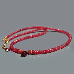 Könnte beinhalten: Eine rote Edelstein-Halskette mit einem goldenen Verschluss und einem tropfenförmigen Anhänger. Die Halskette besteht aus kleinen, facettierten roten Perlen. Der goldene Verschluss und Anhänger verleihen dem Schmuck Eleganz.