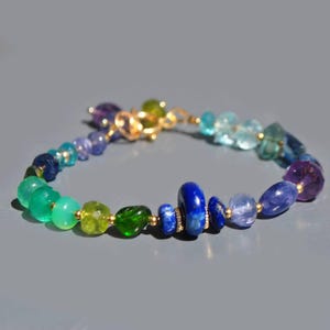 Può includere: Un braccialetto di pietre preziose colorate con una varietà di pietre lucide nelle tonalità del verde, blu, viola e oro. Il braccialetto è infilato su una catena d'oro e ha una chiusura in oro. Le pietre variano per dimensioni e forma.