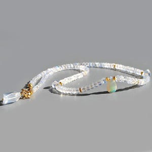 May include: A beaded necklace featuring translucent, faceted moonstone beads with a subtle blue sheen. The strand is accented with small gold-toned spacers and a single, smooth, pear-shaped opal pendant. A clear, faceted crystal charm hangs near the gold clasp.