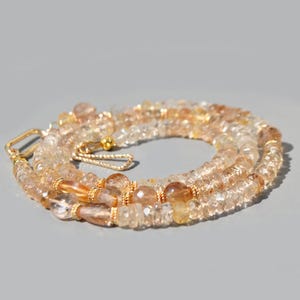 May include: A necklace composed of faceted, translucent light brown and golden citrine beads. The strand features small gold-toned metal spacers and a gold-toned hook-and-eye clasp mechanism, arranged in a circular, coiled formation.