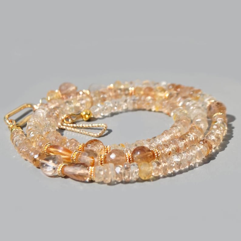 May include: A necklace composed of faceted, translucent light brown and golden citrine beads. The strand features small gold-toned metal spacers and a gold-toned hook-and-eye clasp mechanism, arranged in a circular, coiled formation.