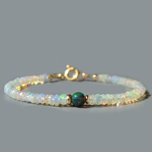 Op de afbeelding: Een delicate armband gemaakt met kleine witte opaal kralen en een enkele grotere groene opaal kraal in het midden. De armband is beveiligd met een gouden sluiting.