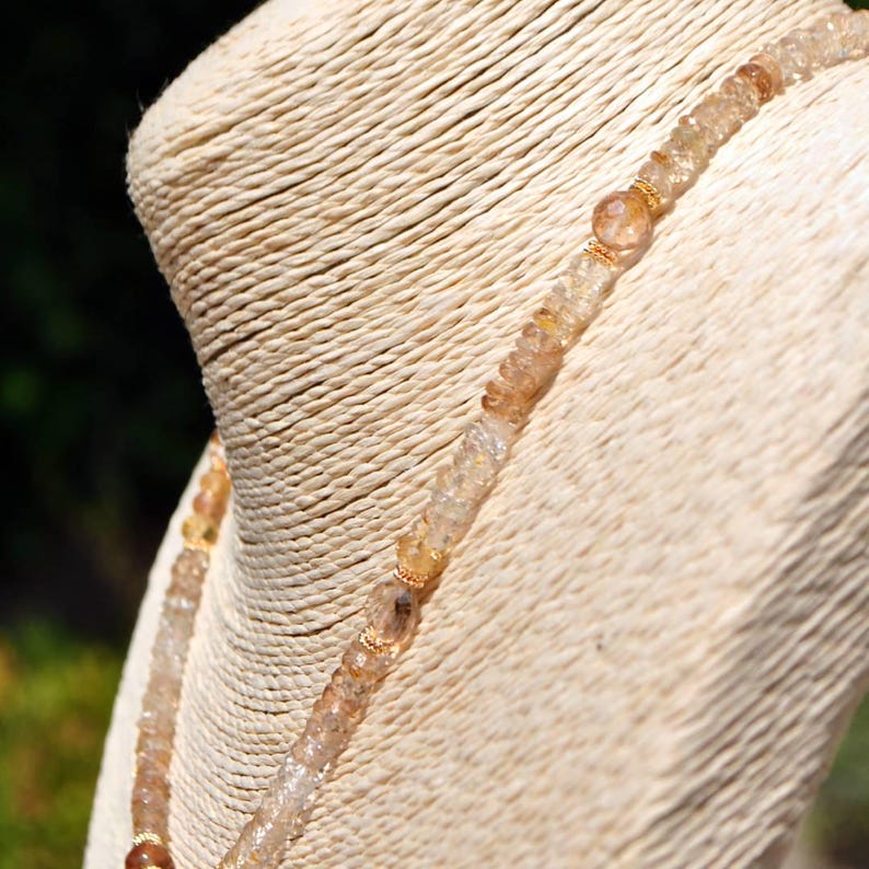 May include: A beaded necklace featuring faceted, translucent amber-toned gemstones interspersed with small, round, metallic gold-colored spacers. The beads vary slightly in shade and clarity, creating a textured, shimmering appearance along the strand.
