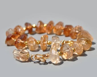 May include: A bracelet featuring faceted, translucent amber-colored gemstone beads interspersed with small, round, polished gold-tone metal beads. The bracelet is arranged in a circular shape and includes a gold-tone metal lobster claw clasp.