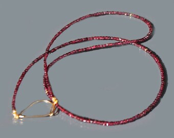 Op de afbeelding: Een lange ketting gemaakt van kleine, donkerrode kralen met een gouden hanger in de vorm van een traan.