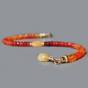 May include: A gemstone bracelet with a colour gradient of orange and red beads. The bracelet has a gold clasp and two oval opal stones. The gemstones transition from deep red to a vibrant orange hue.