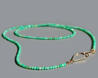 May include: A delicate necklace featuring small, round, light green beads. The beads are strung together, forming a long strand that curves gracefully. A gold-colored clasp secures the necklace, adding a touch of elegance. The necklace is displayed against a gray background.
