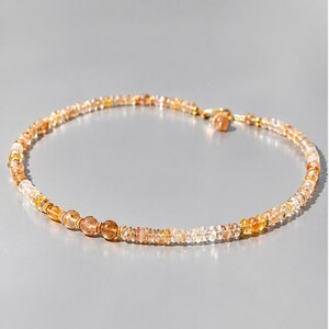 May include: A beaded necklace featuring a series of small, translucent, amber-toned gemstone beads in varying shades of yellow and orange. The necklace includes a gold-toned metal clasp and a small dangling charm at the closure point.