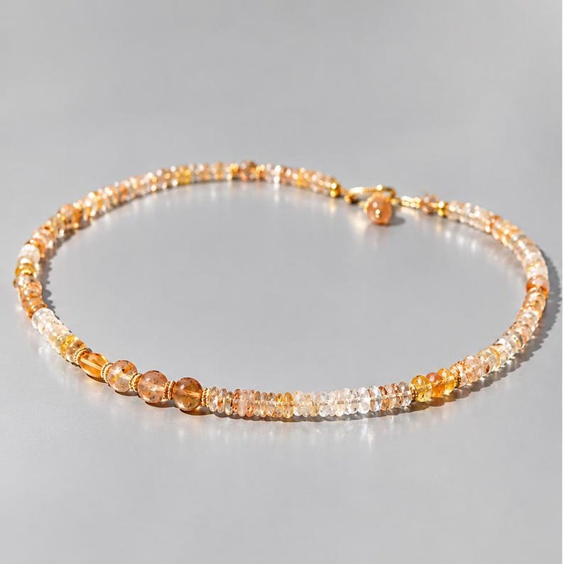 May include: A beaded necklace featuring a series of small, translucent, amber-toned gemstone beads in varying shades of yellow and orange. The necklace includes a gold-toned metal clasp and a small dangling charm at the closure point.