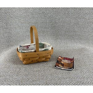 May include: A rectangular woven wooden basket with a single upright handle and a removable fabric liner featuring a floral pattern. Beside the basket lies a small rectangular paper tag with the text Classic Baskets and Longaberger printed on it.