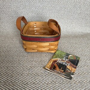 May include: A square woven wooden basket with two tan leather handles and a dark red accent band around the top rim. Beside the basket lies a small informational card featuring the text 1999 Father's Day Tee Basket Longaberger.