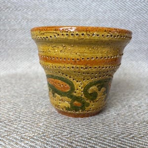Puede incluir: Una maceta de cerámica hecha a mano con un esmalte amarillo y naranja. La maceta presenta un patrón decorativo de diseños florales verdes y naranjas y bandas horizontales de pequeños agujeros. El borde es de un tono naranja más oscuro.