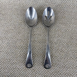Op de afbeelding: Twee zilverkleurige metalen lepels met een gepolijste afwerking. De ene lepel heeft een dichte ovale kom, terwijl de andere een schuimspaan is met vijf langwerpige openingen. Beide handvatten eindigen in een decoratief schelpmotief aan de basis.
