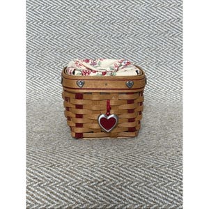 May include: A small, square woven basket with a red and natural wood color scheme. The basket features a red heart-shaped charm and a floral patterned fabric lining the interior. Decorative metal hearts adorn the top rim.