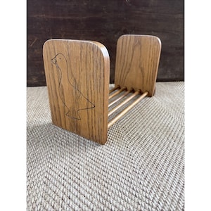 Puede incluir: Un estante para libros de madera con dos paneles verticales conectados por cuatro varillas horizontales. El panel frontal muestra un dibujo lineal negro de un ganso. La madera tiene una veta visible y un acabado en tono marrón claro.