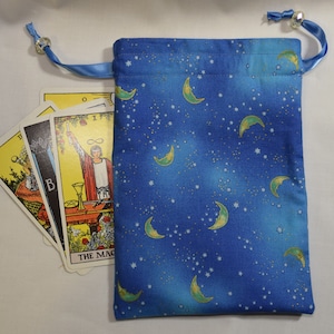 Könnte beinhalten: Blaue Stofftasche mit Kordelzug und einem Muster aus Halbmonden und Sternen. Die Tasche ist gefüllt mit Tarotkarten, darunter die Karte des Magiers.