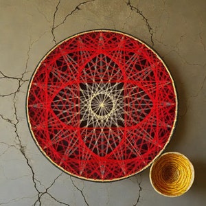 Könnte beinhalten: Ein rundes String-Art-Design in Rot und Weiß. Das Design besteht aus vielen sich kreuzenden Linien, die ein geometrisches Muster bilden. Die String Art ist auf einem Holzrahmen montiert.