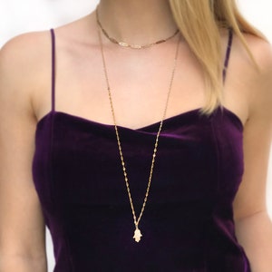 Könnte beinhalten: Eine goldene Kettenkette mit einem Hamsa-Hand-Anhänger. Die Kette ist über eine zweite, kürzere Kette gelegt. Das Model trägt ein Oberteil aus lila Samt.