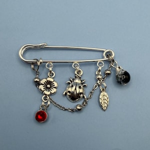 Puede incluir: Un broche de imperdible plateado con colgantes. Los colgantes incluyen una flor, una mariquita, una hoja y una gema roja. El broche está sobre un fondo azul claro.