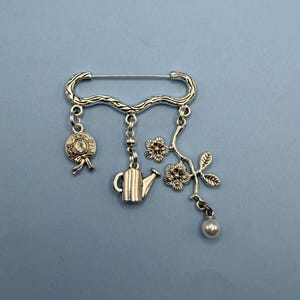 May include: Silver-toned brooch featuring a decorative bar with dangling charms. Charms include a bow, a teapot, flowers, and a faux pearl. The brooch is set against a solid blue background.