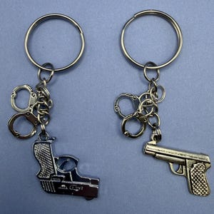 May include: Two keychains with metal charms. One keychain features a silver gun charm and a pair of handcuffs. The other keychain has a gold gun charm and a pair of handcuffs. Both keychains have a silver ring.