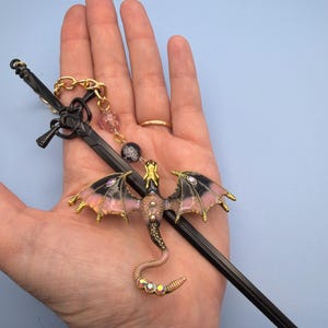 May include: A black sword-shaped hair stick with a gold chain and a dragon charm. The dragon has pink and black wings, a gold head, and a jeweled tail. The charm is attached to the hair stick with a gold chain and pink and black beads.