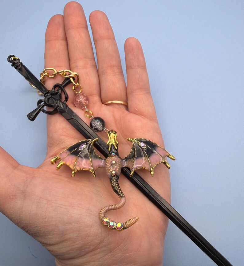 May include: A black sword-shaped hair stick with a gold chain and a dragon charm. The dragon has pink and black wings, a gold head, and a jeweled tail. The charm is attached to the hair stick with a gold chain and pink and black beads.