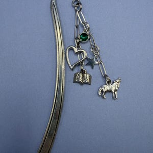 May include: Silver metal bookmark with a curved top and dangling charms. Charms include a heart, a book, a star, a green gem, and a howling wolf. The bookmark is on a light blue background.