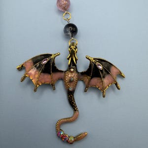 May include: A gold-toned dragon pendant with black and pink wings, adorned with purple and iridescent gemstones. The dragon's body is a mix of pink and brown, with a curled tail. The pendant hangs from a gold chain with pink and black beads.